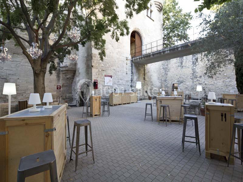 Cour extérieure aménagée avec des tables hautes en bois, des tabourets métalliques et des lampes, entourée de murs en pierre.