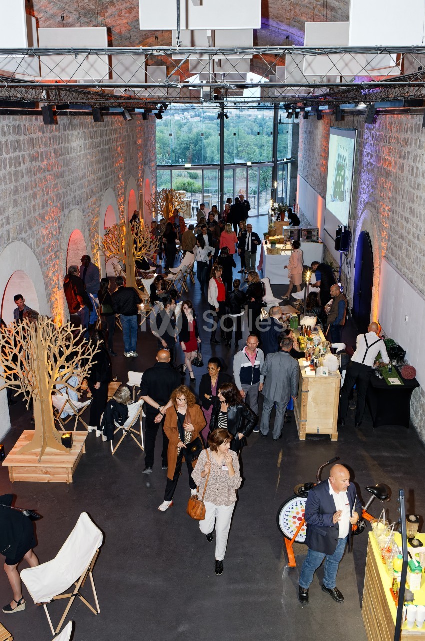 Salle animée avec des invités discutant autour de stands, dans un espace lumineux aux murs en pierre et grandes fenêtres.