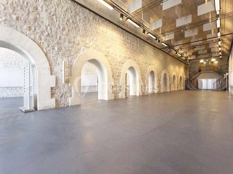 Salle spacieuse avec murs en pierre, arches, sol lisse et plafond équipé de luminaires modernes.