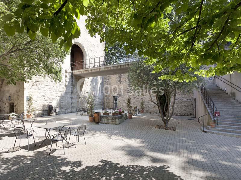 Cour pavée avec des tables et chaises en métal, entourée de murs en pierre, arbres et un escalier sur la droite.