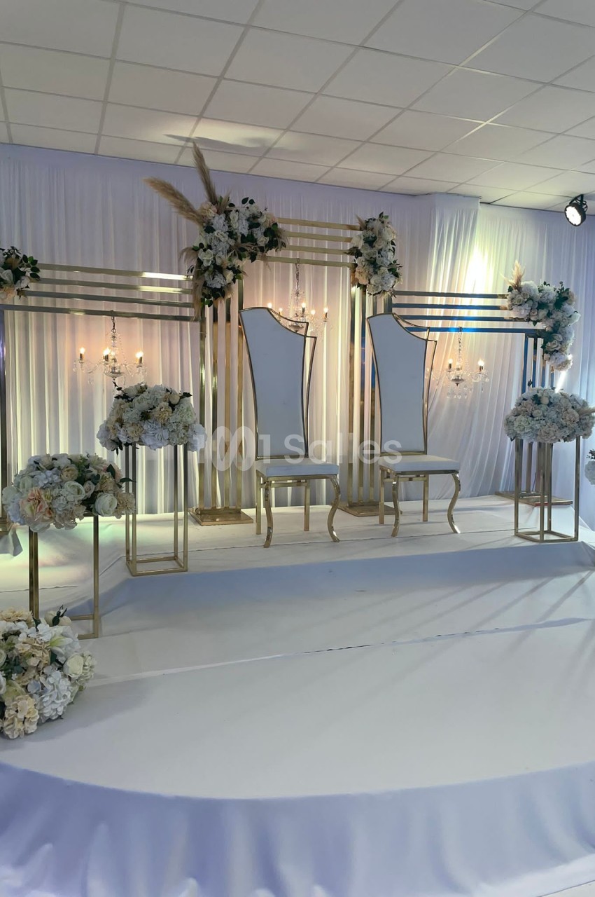 Deux chaises élégantes sur une scène décorée de fleurs blanches et dorées, avec un fond en tissu plissé.
