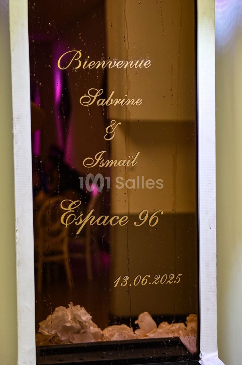 Texte doré sur une vitre indiquant ’Bienvenue Sabrine & Ismail, Espace 96, 13.06.2025’, avec des fleurs blanches en bas.