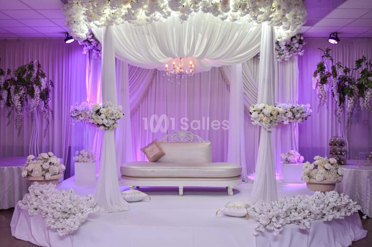 Décor de mariage avec canapé blanc sous une arche drapée de tissu blanc, entourée de fleurs et éclairée en violet.