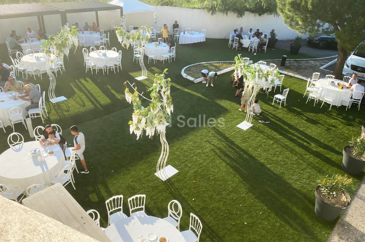 Jardin aménagé pour un événement avec des tables rondes, des chaises blanches et des décorations florales suspendues.