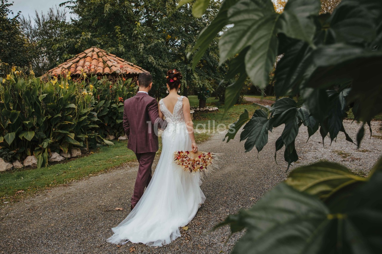 Un couple en tenue de mariage marche sur un chemin bordé de verdure, près d'une petite cabane en pierre.