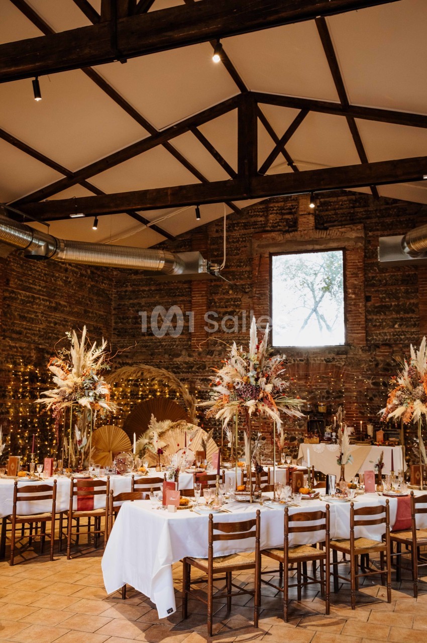 Salle de réception rustique avec tables décorées de nappes blanches, fleurs séchées et guirlandes lumineuses.