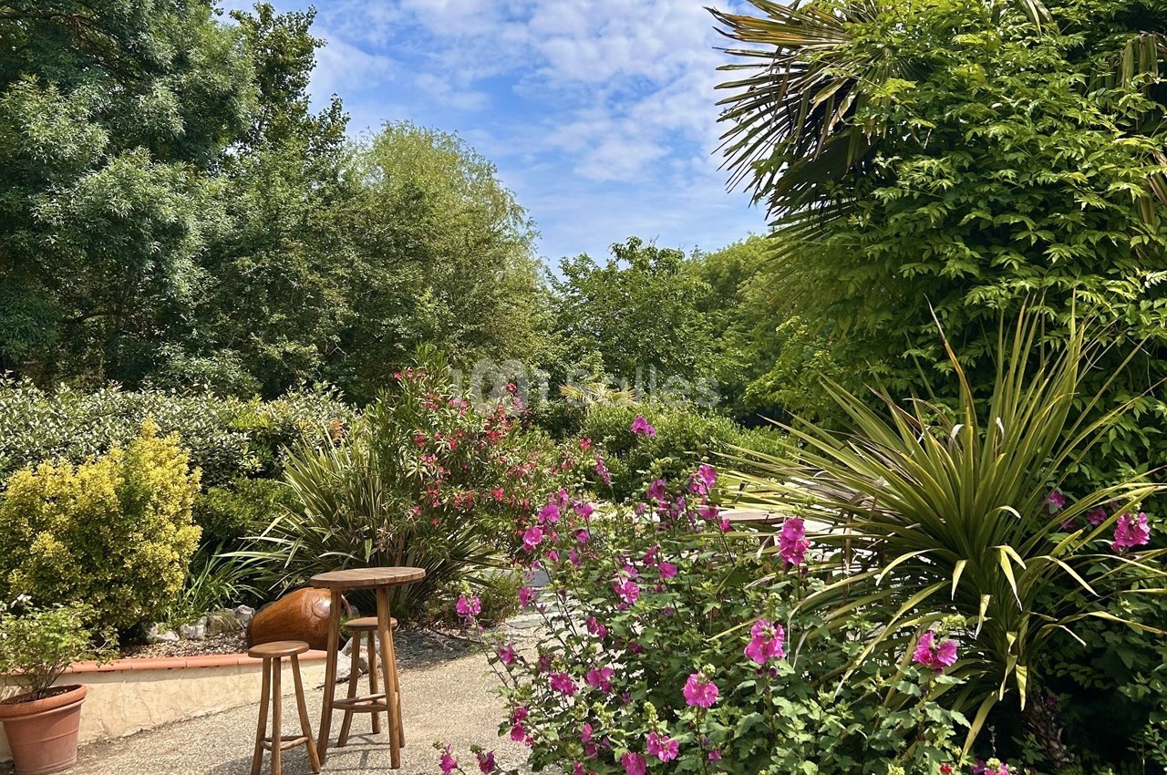 Jardin verdoyant avec fleurs roses, arbustes et palmiers sous un ciel partiellement nuageux.