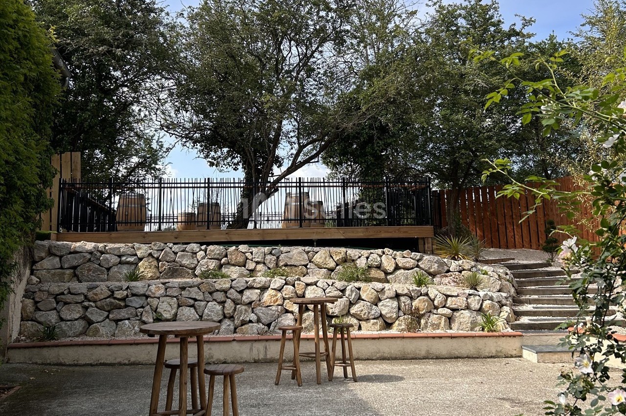Terrasse surélevée avec tables et chaises, entourée d'arbres et de pierres, vue depuis une cour avec tabourets en bois.