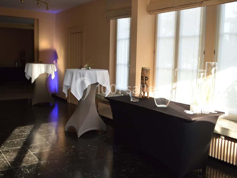 Salle lumineuse avec tables hautes drapées de nappes blanches et une table basse noire près de grandes fenêtres.