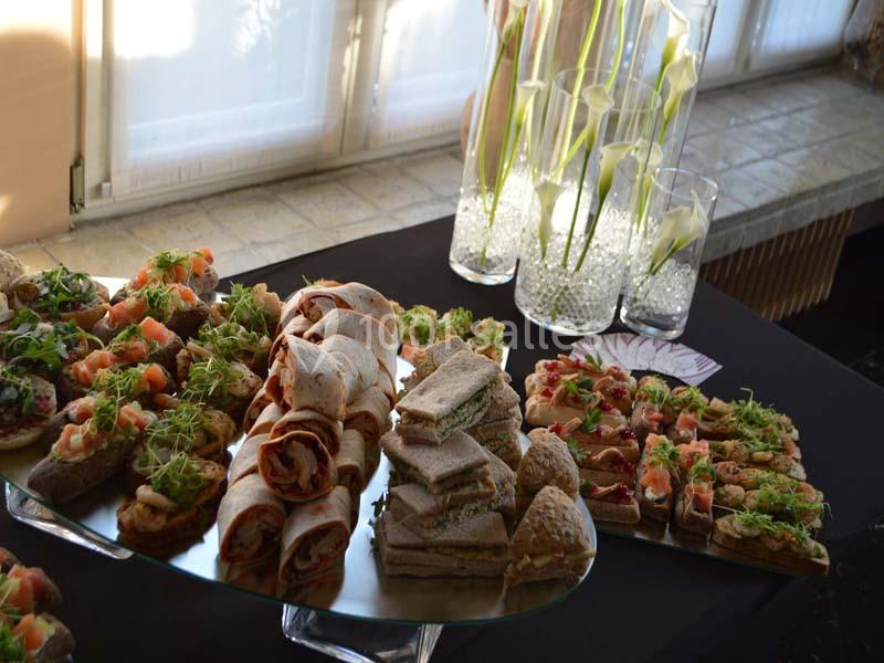 Plateau de canapés variés, wraps et sandwichs disposés sur une table noire près de vases avec des fleurs blanches.