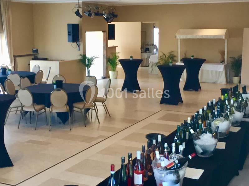 Salle de réception avec tables rondes dressées, mange-debout noirs et un buffet de bouteilles de vin sur glace.
