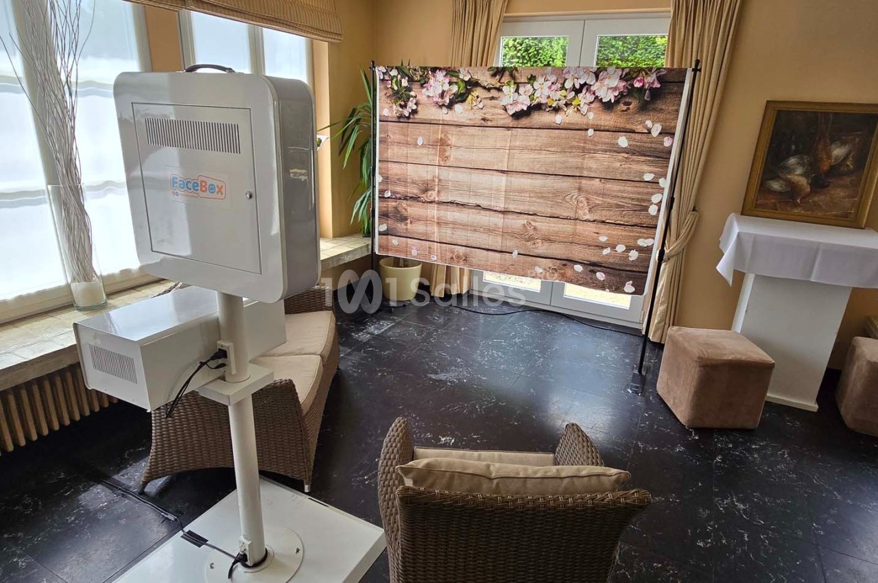 Cabine photo installée dans une pièce lumineuse avec un fond en bois décoré de fleurs et des fauteuils en osier.