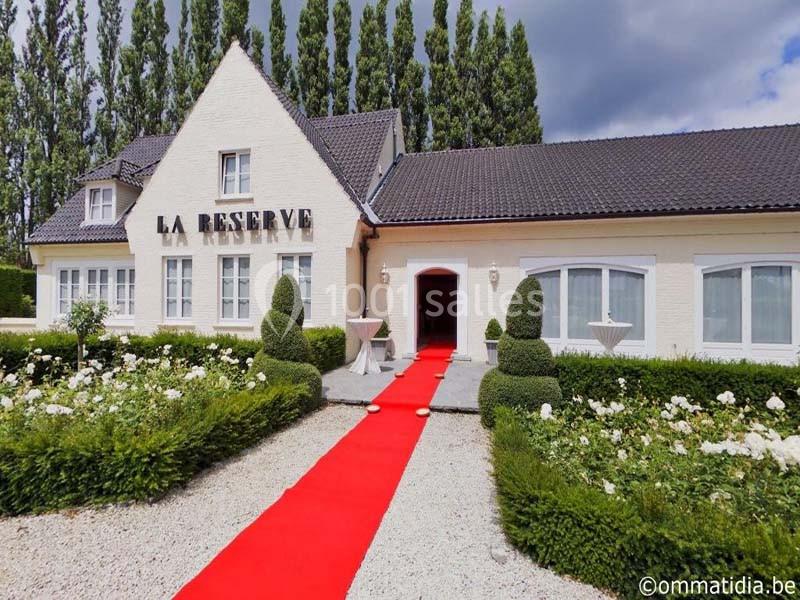 Façade d'un bâtiment blanc avec une entrée ornée d'un tapis rouge, entourée de buissons taillés et de fleurs blanches.