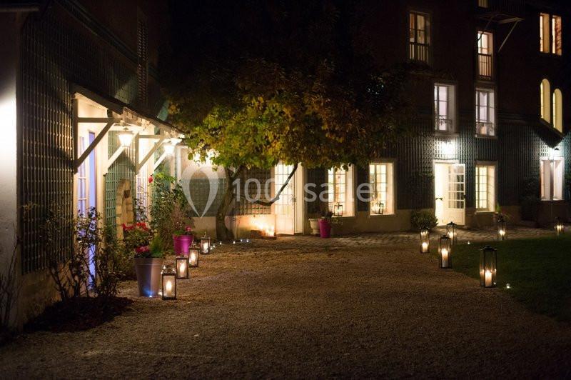 Cour éclairée en soirée avec des lanternes au sol, un arbre central et une façade de bâtiment aux fenêtres illuminées.