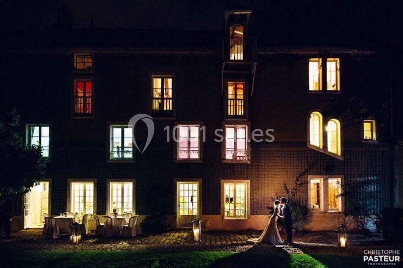 Un couple se tient enlacé devant une grande maison éclairée de nuit, avec des lanternes posées sur le sol.