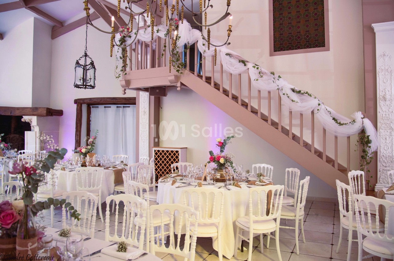 Salle de réception décorée avec des tables rondes, des chaises blanches et un escalier orné de guirlandes blanches.