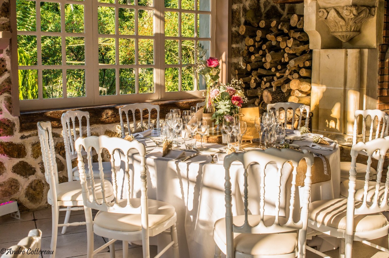 Table ronde élégamment dressée avec des chaises blanches, décorée de fleurs, près d'une fenêtre laissant entrer la lumière…