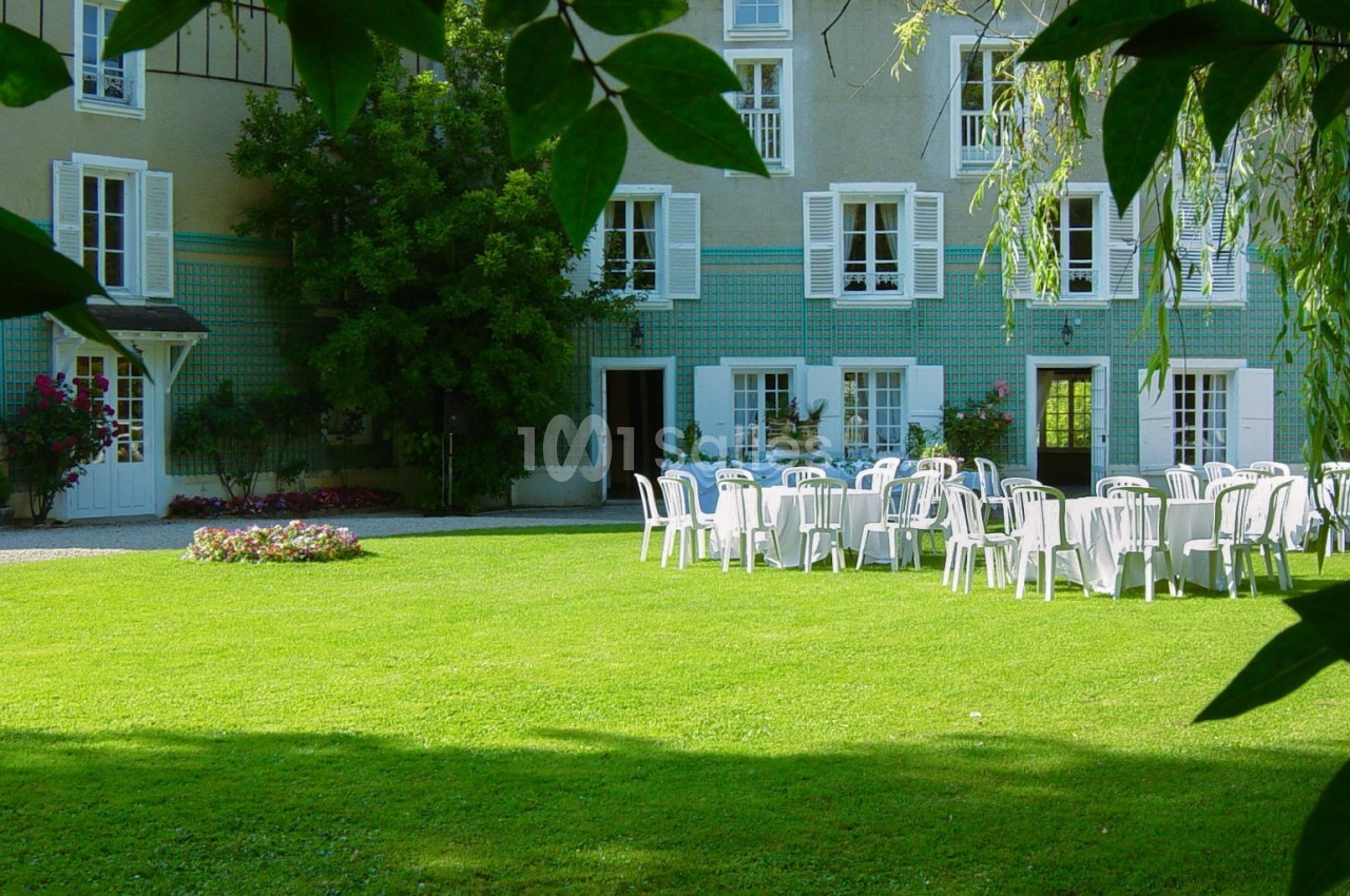 Chaises blanches disposées autour de tables sur une pelouse devant une grande maison avec volets blancs.