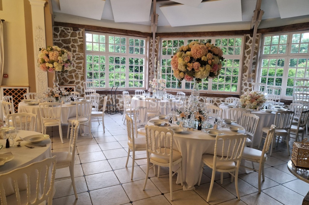 Salle de réception élégante avec tables rondes dressées, chaises blanches et centres de table fleuris.