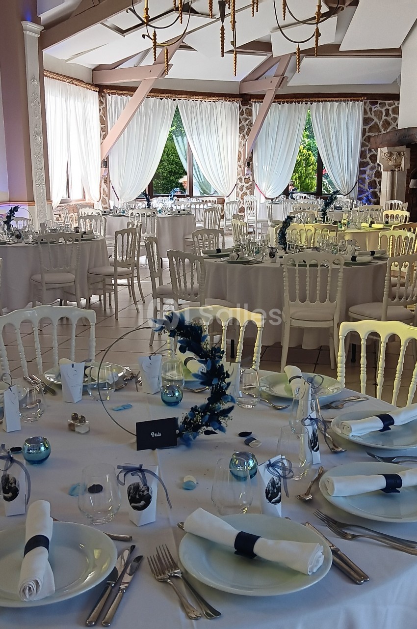 Salle de réception décorée avec des tables rondes dressées, nappes blanches et centres de table floraux bleus.