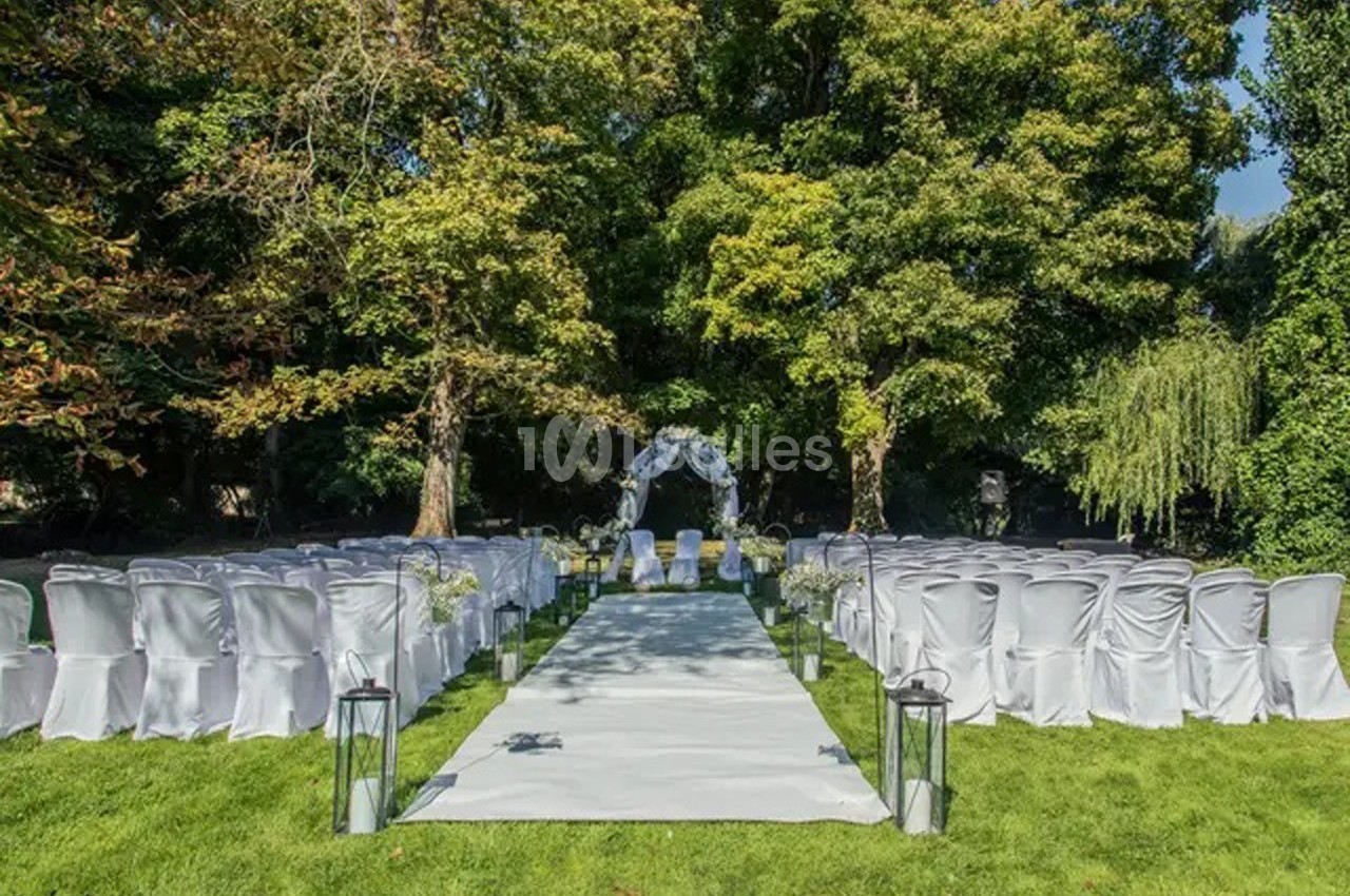 Allée centrale décorée pour une cérémonie de mariage en extérieur, entourée de chaises blanches et d'arbres verdoyants.
