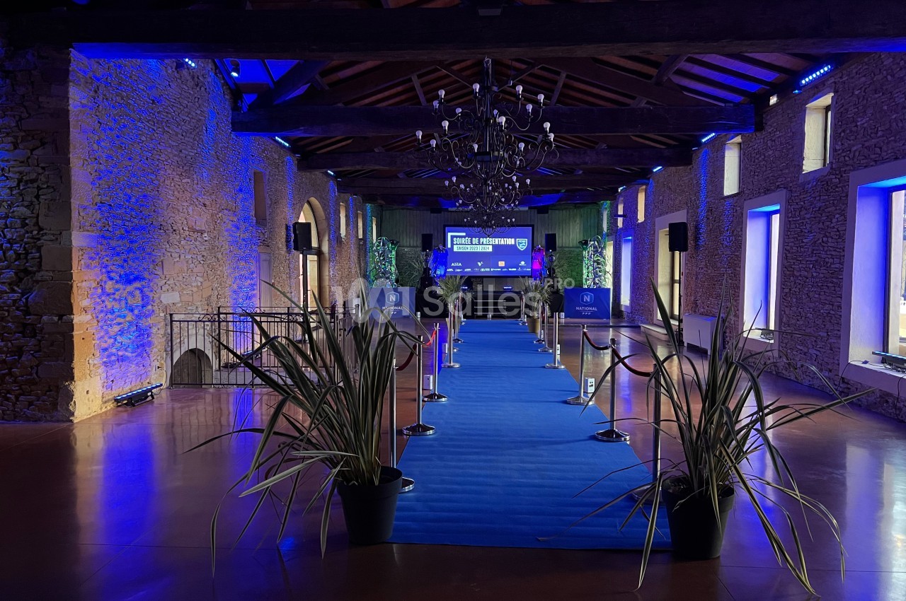 Salle en pierre avec un tapis bleu menant à une scène éclairée, décorée de plantes et de luminaires suspendus.