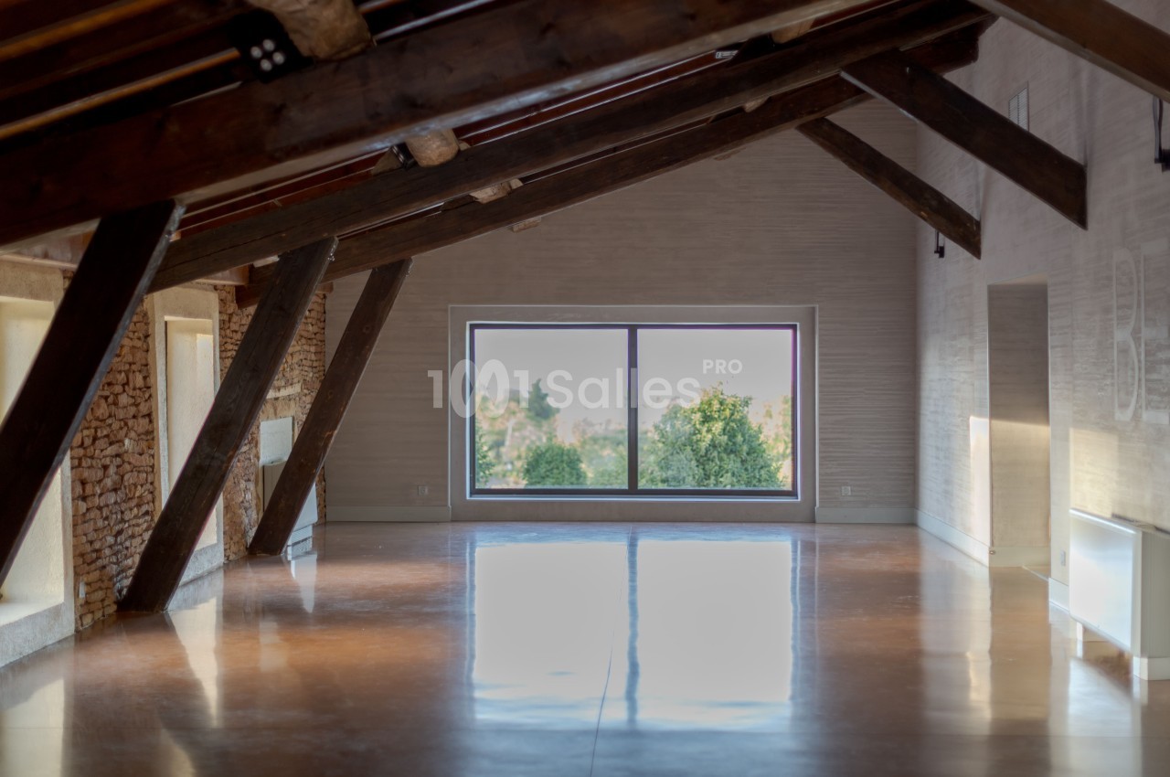 Vue intérieure d'une pièce mansardée lumineuse avec poutres apparentes, sol en bois et une grande fenêtre donnant sur des…