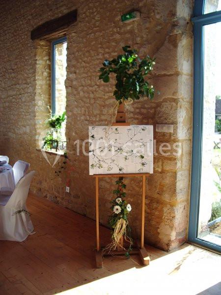 Plan de table décoré de fleurs et de feuillage, posé sur un chevalet en bois dans une salle en pierre lumineuse.