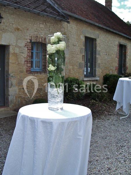 Vase cylindrique avec des roses blanches sur une table ronde drapée de blanc, devant une maison en pierre.