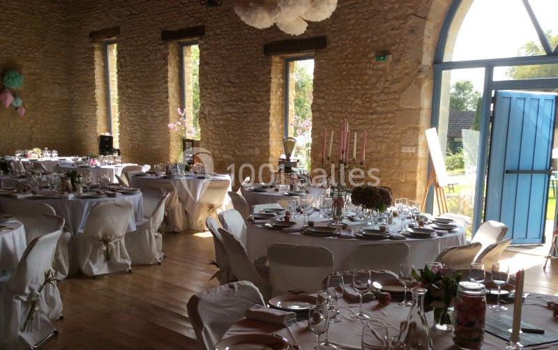 Salle de réception en pierre avec tables rondes dressées pour un événement, lumière naturelle entrant par une grande porte…