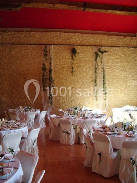 Salle de réception avec murs en pierre, tables rondes dressées et chaises blanches décorées de feuillage.