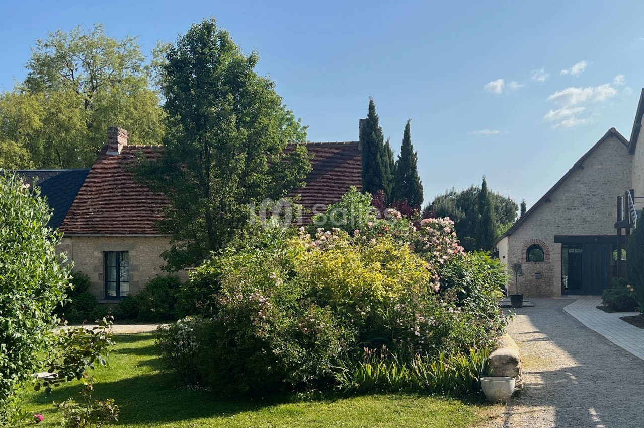 Cour d'une maison en pierre avec jardin fleuri, allée pavée et arbres sous un ciel dégagé.