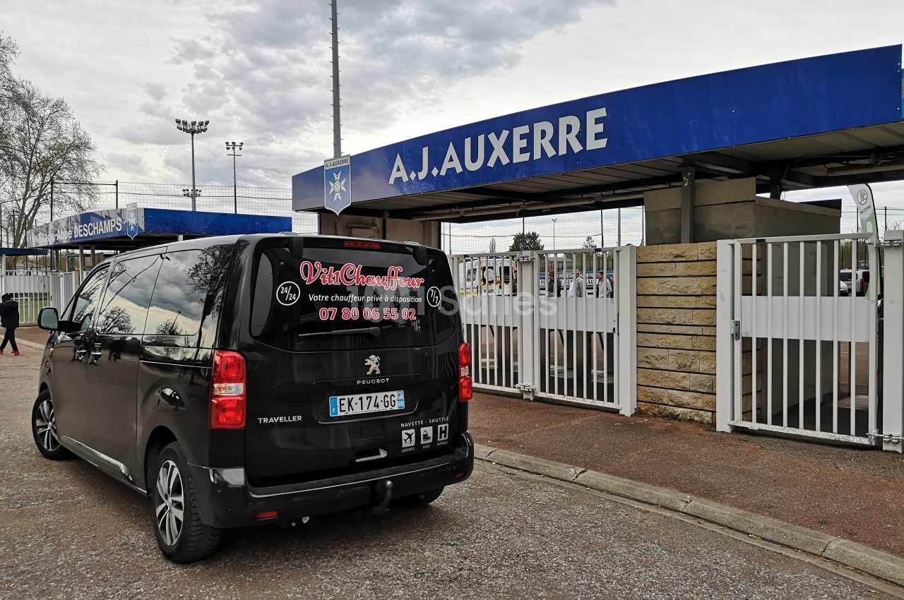 Un minivan noir stationné devant l'entrée principale du stade de l'A.J. Auxerre par temps nuageux.