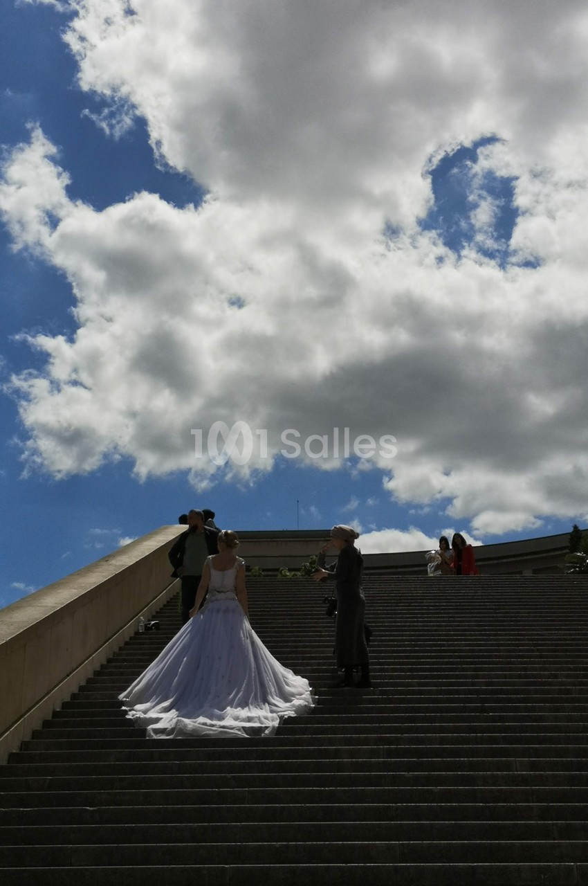 Un couple en tenue de mariage pose sur un large escalier extérieur sous un ciel partiellement nuageux.