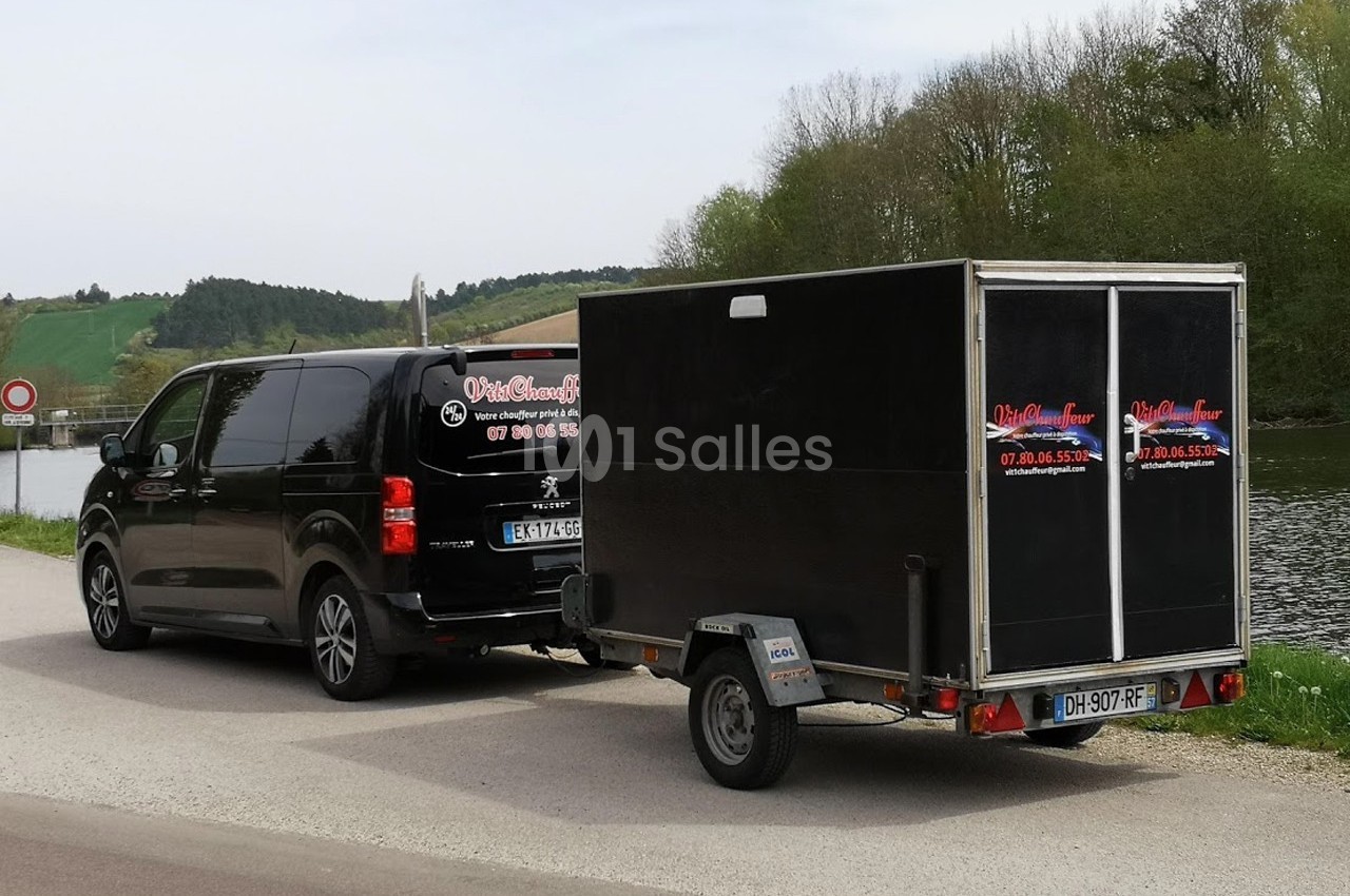 Un van noir Peugeot tractant une remorque noire sur une route près d'un cours d'eau et d'une zone boisée.