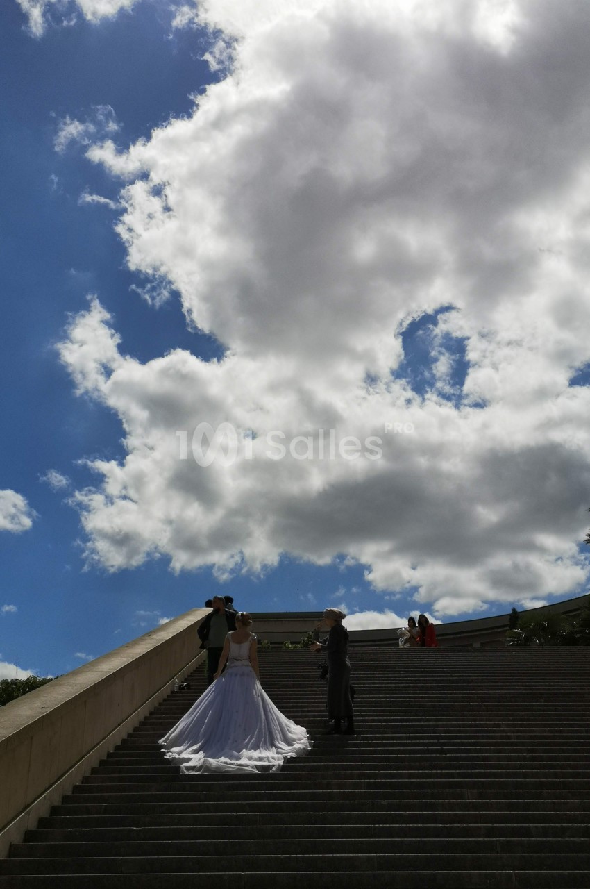 Un couple en tenue de mariage pose sur un large escalier sous un ciel partiellement nuageux.