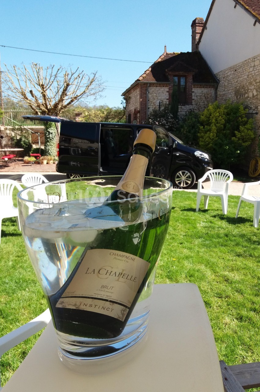 Bouteille de champagne dans un seau à glace sur une table, avec jardin, chaises blanches et véhicule en arrière-plan.