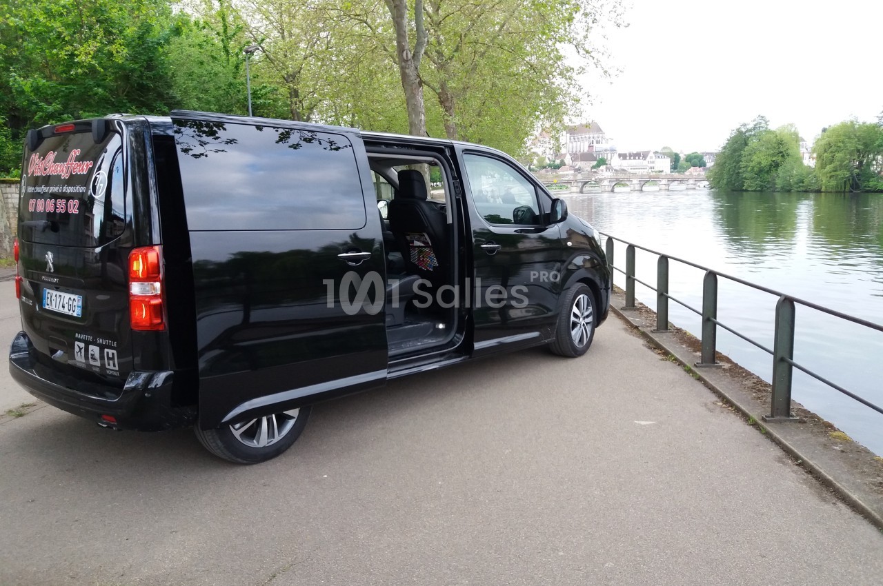 Un van noir stationné près d'un chemin longeant une rivière bordée d'arbres, avec une porte latérale ouverte.