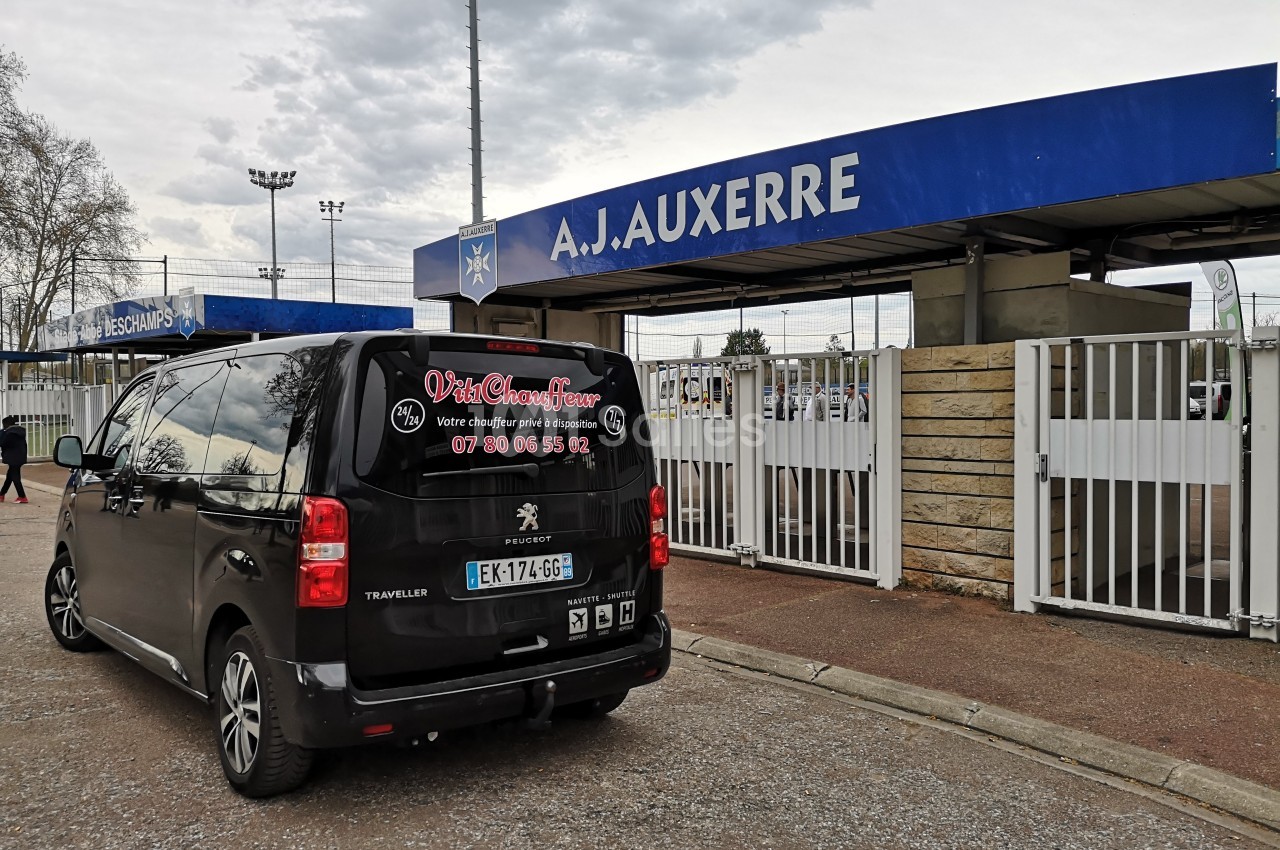 Un van noir stationné devant l'entrée principale du stade de l'AJ Auxerre par temps nuageux.