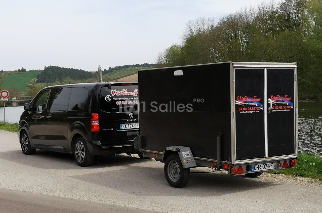 Un van noir Peugeot tractant une remorque noire sur une route près d'un cours d'eau et d'une zone boisée.