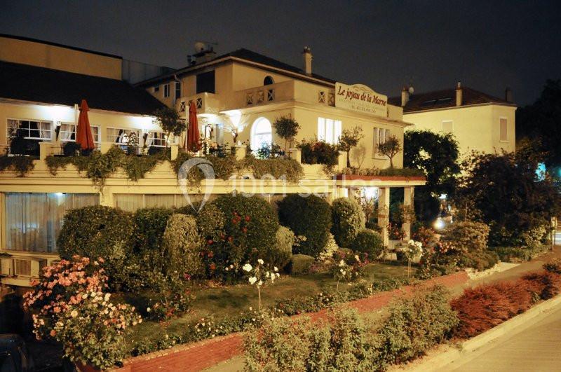 Façade d'un restaurant entouré d'un jardin fleuri, éclairé la nuit dans un quartier résidentiel.