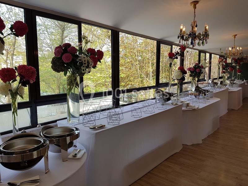 Salle lumineuse avec tables de buffet dressées, décorées de grandes compositions florales et vue sur un jardin.