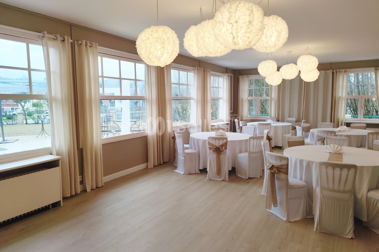 Salle lumineuse avec grandes fenêtres, tables rondes dressées de nappes blanches et chaises décorées de nœuds beiges.
