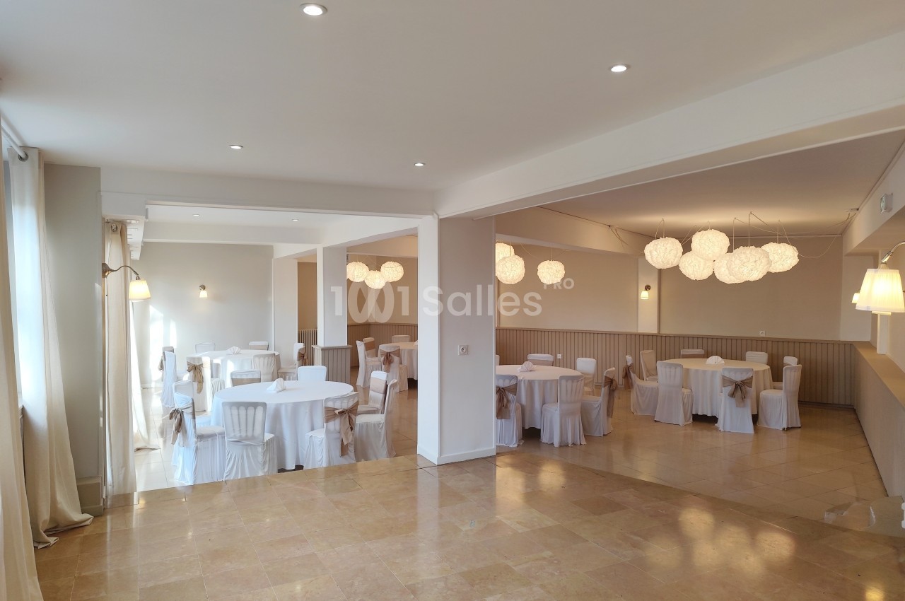 Salle lumineuse avec tables rondes dressées de nappes blanches, chaises assorties et suspensions décoratives modernes.