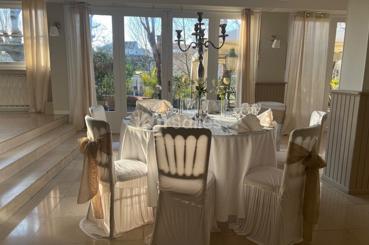 Table ronde élégamment dressée avec nappes blanches, chaises décorées et chandelier, dans une salle lumineuse avec vue sur…