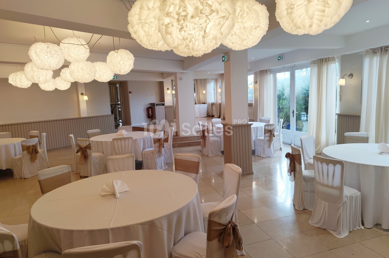 Salle de réception lumineuse avec tables rondes dressées de nappes blanches et décorations suspendues en papier.