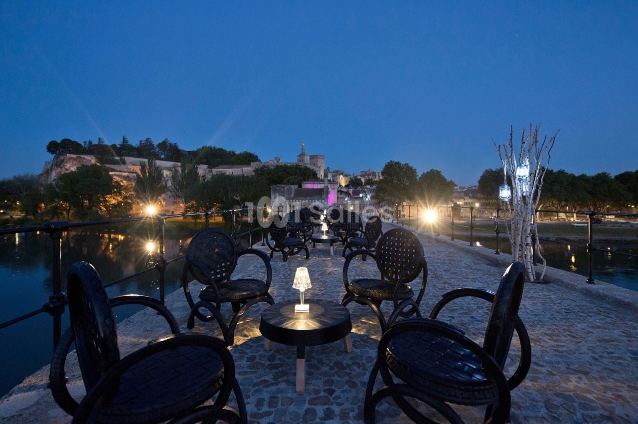 Terrasse extérieure avec tables et chaises noires, éclairée par des lampes, vue sur un fleuve et des bâtiments illuminés.