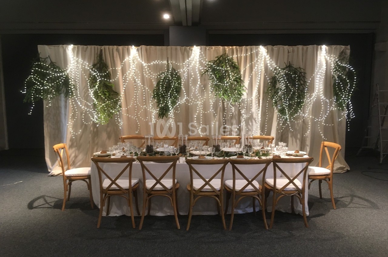 Table décorée pour un événement, entourée de chaises en bois, avec un fond de rideaux, guirlandes lumineuses et feuillages.