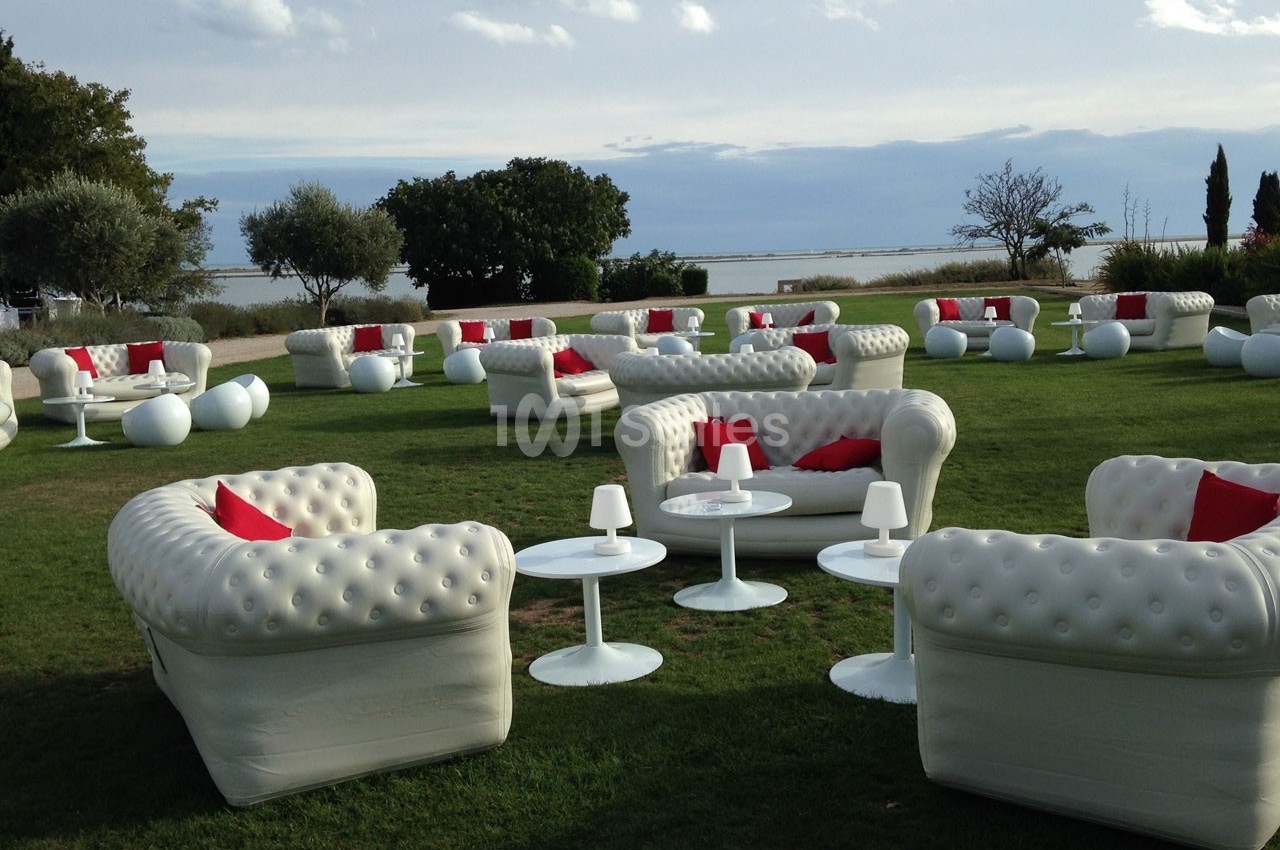 Mobilier de salon blanc avec coussins rouges disposé sur une pelouse en extérieur, près d'un plan d'eau.