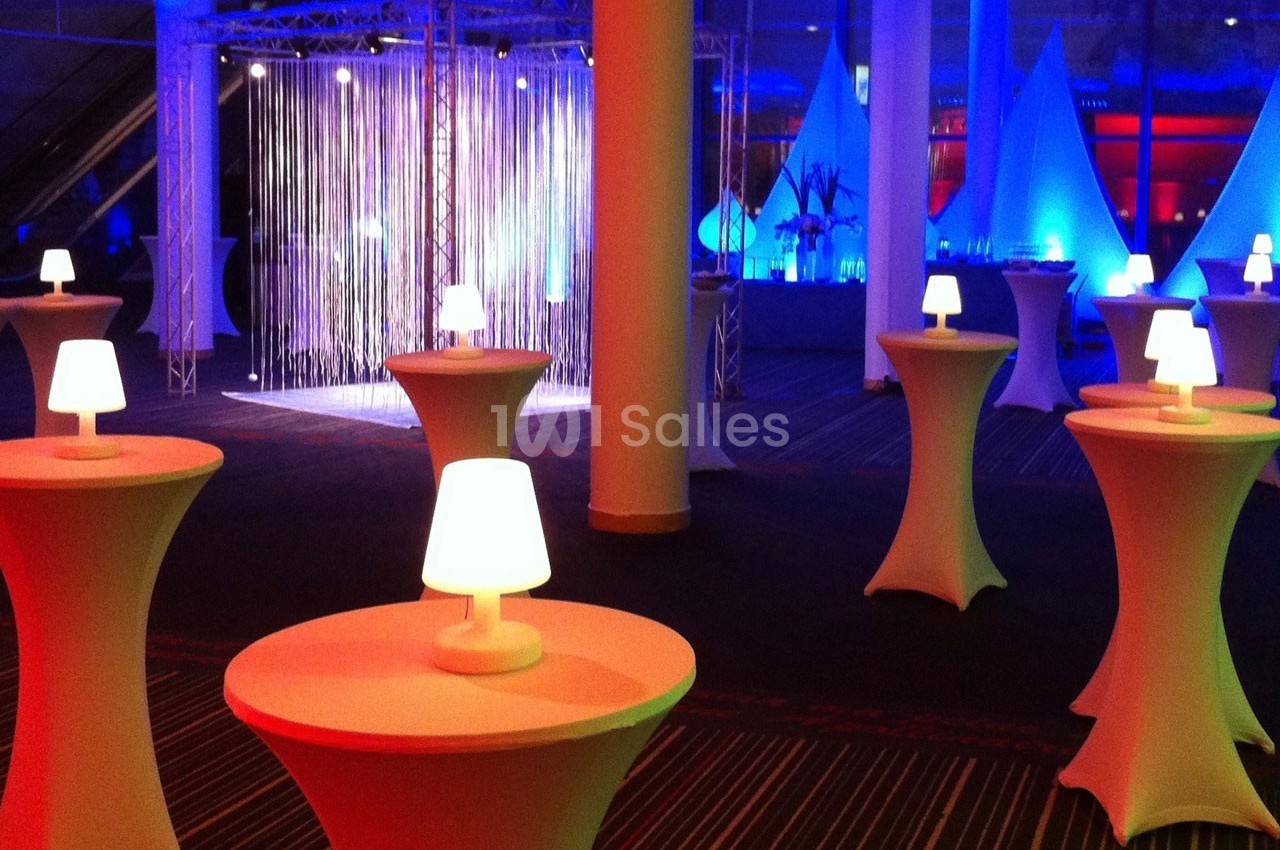 Salle décorée pour un événement avec des tables hautes éclairées et un rideau lumineux en arrière-plan.